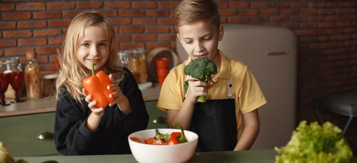 Cute girl and boy with fresh pepper and broccoli in hands standing near desk with assorted vegetables and salad bowl during vegetarian meal preparation