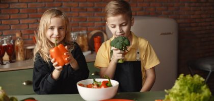 Cute girl and boy with fresh pepper and broccoli in hands standing near desk with assorted vegetables and salad bowl during vegetarian meal preparation