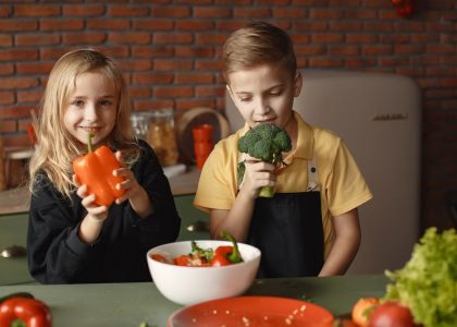 Cute girl and boy with fresh pepper and broccoli in hands standing near desk with assorted vegetables and salad bowl during vegetarian meal preparation